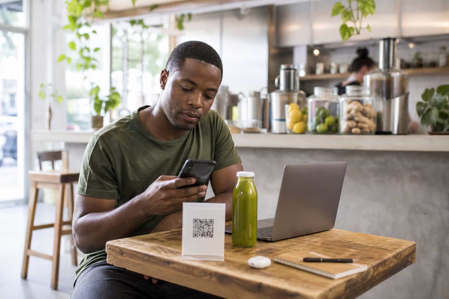 Jovem cliente negro do sexo masculino perto dos trinta anos escaneando um QR code impresso em uma mesa de madeira dentro de uma juice bar iluminada, um notebook aberto e uma garrafa de suco prensado a frio ao lado dele, luz natural suave
