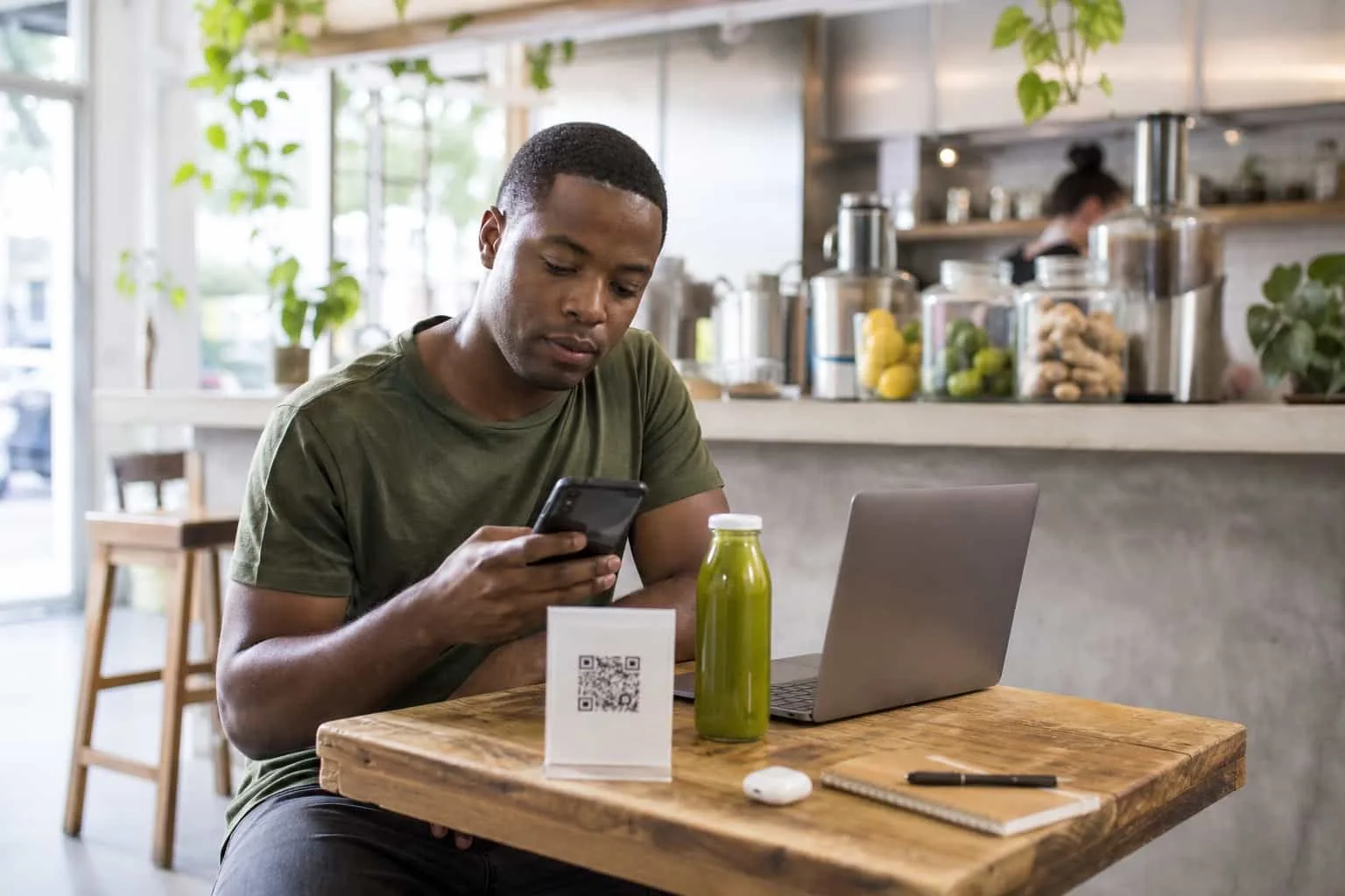 Cliente joven escaneando un código QR impreso sobre una mesa de madera dentro de una juguería luminosa, con una laptop abierta y una botella de jugo prensado en frío al lado, luz natural suave
