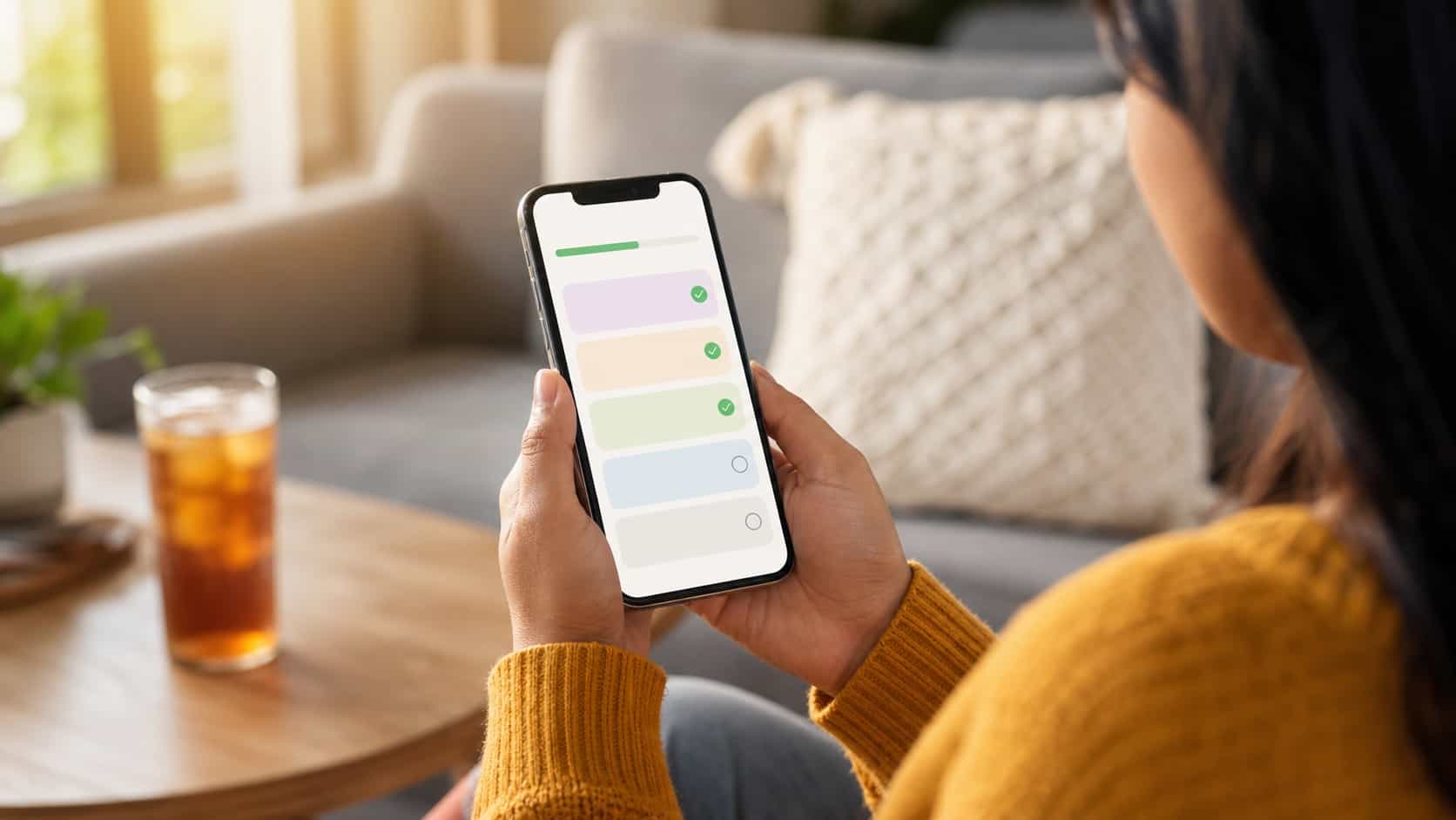 Hands holding a phone showing an abstract task list with progress checkmarks on a couch in afternoon light