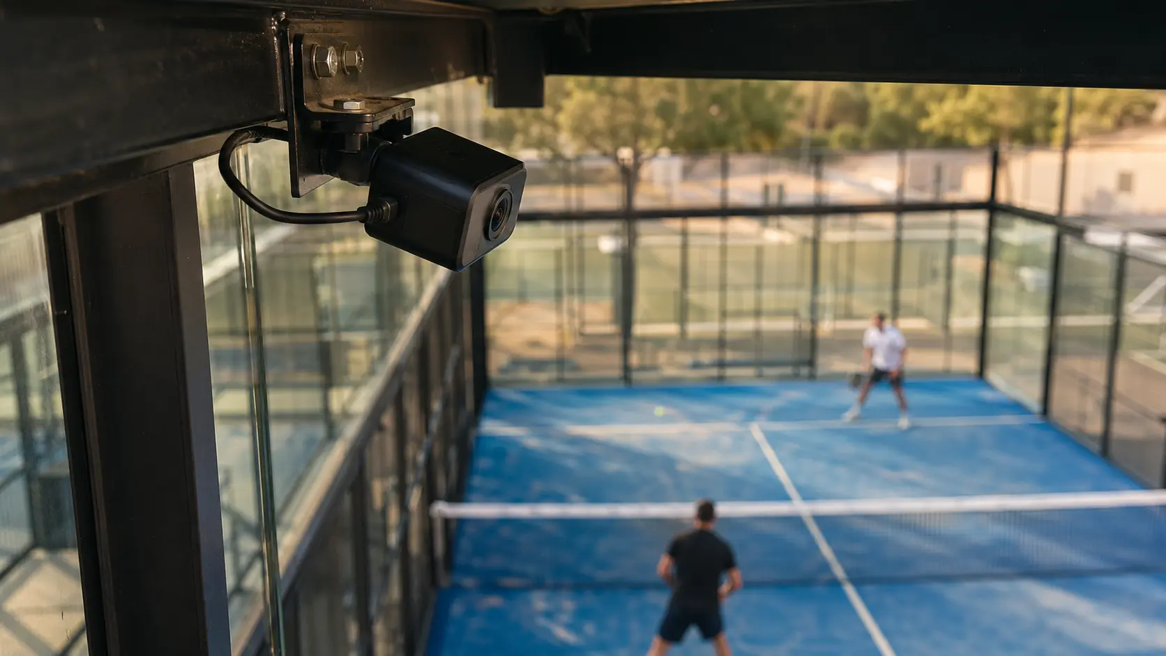Cámara pequeña instalada en alto en la esquina de una cancha cerrada, con la red y el piso visibles abajo