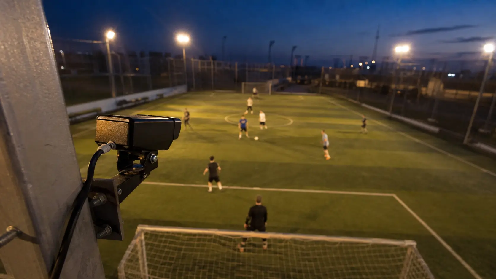 Cámara fija montada detrás del arco de una cancha de fútbol 5 al atardecer, partido en curso bajo los reflectores