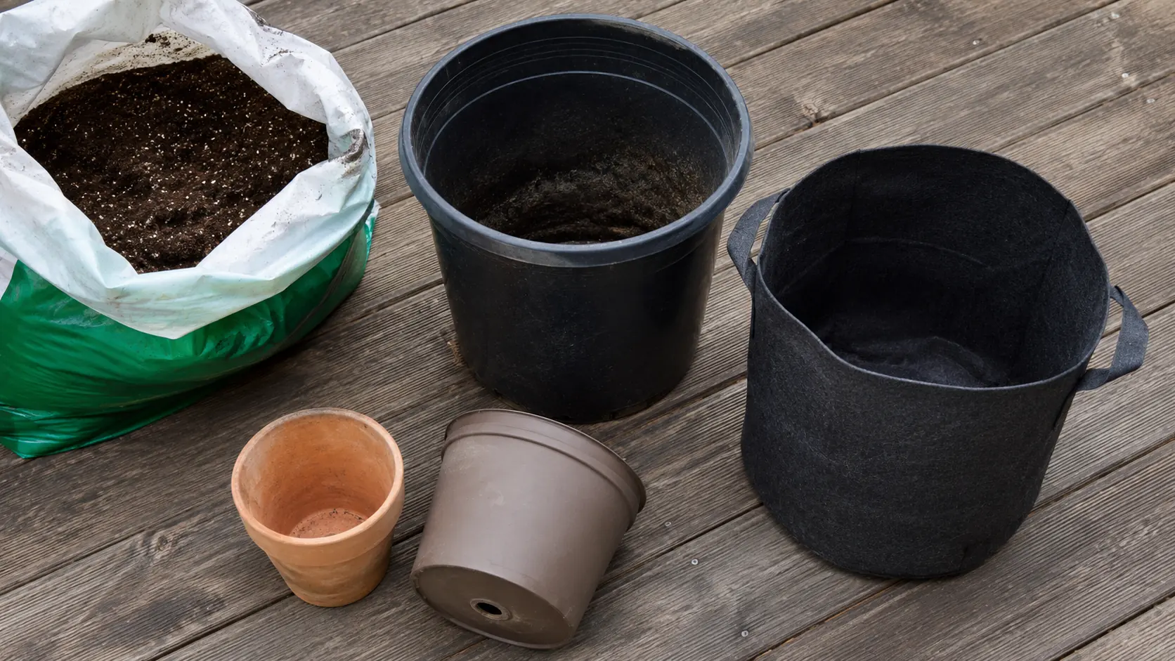 Macetas de jardín vacías alineadas en un deck de madera para comparar tamaños: maceta pequeña de barro, mediana de plástico, grande de 20 litros y bolsa de cultivo de tela, agujero de drenaje visible en una maceta volcada