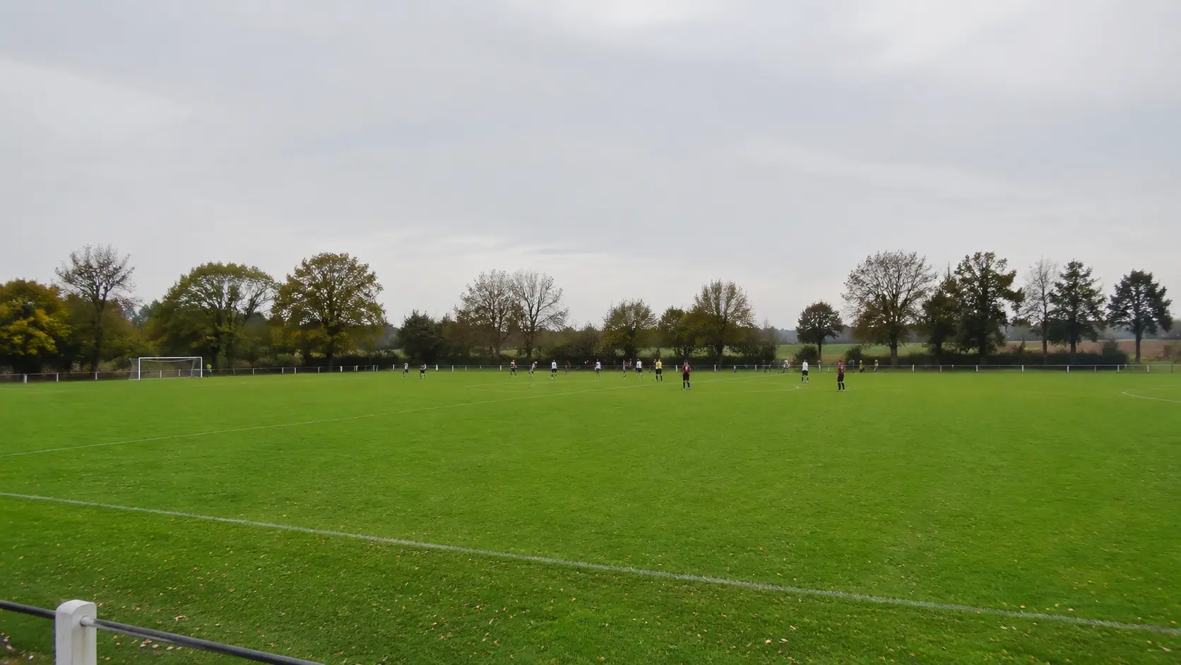 Vista abierta desde el borde de una cancha amateur donde los jugadores se ven pequeños y difíciles de distinguir