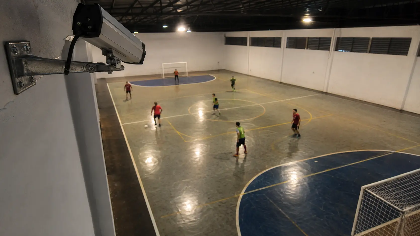 Cámara montada en alto en la esquina de una cancha de futsal techada, partido en juego abajo