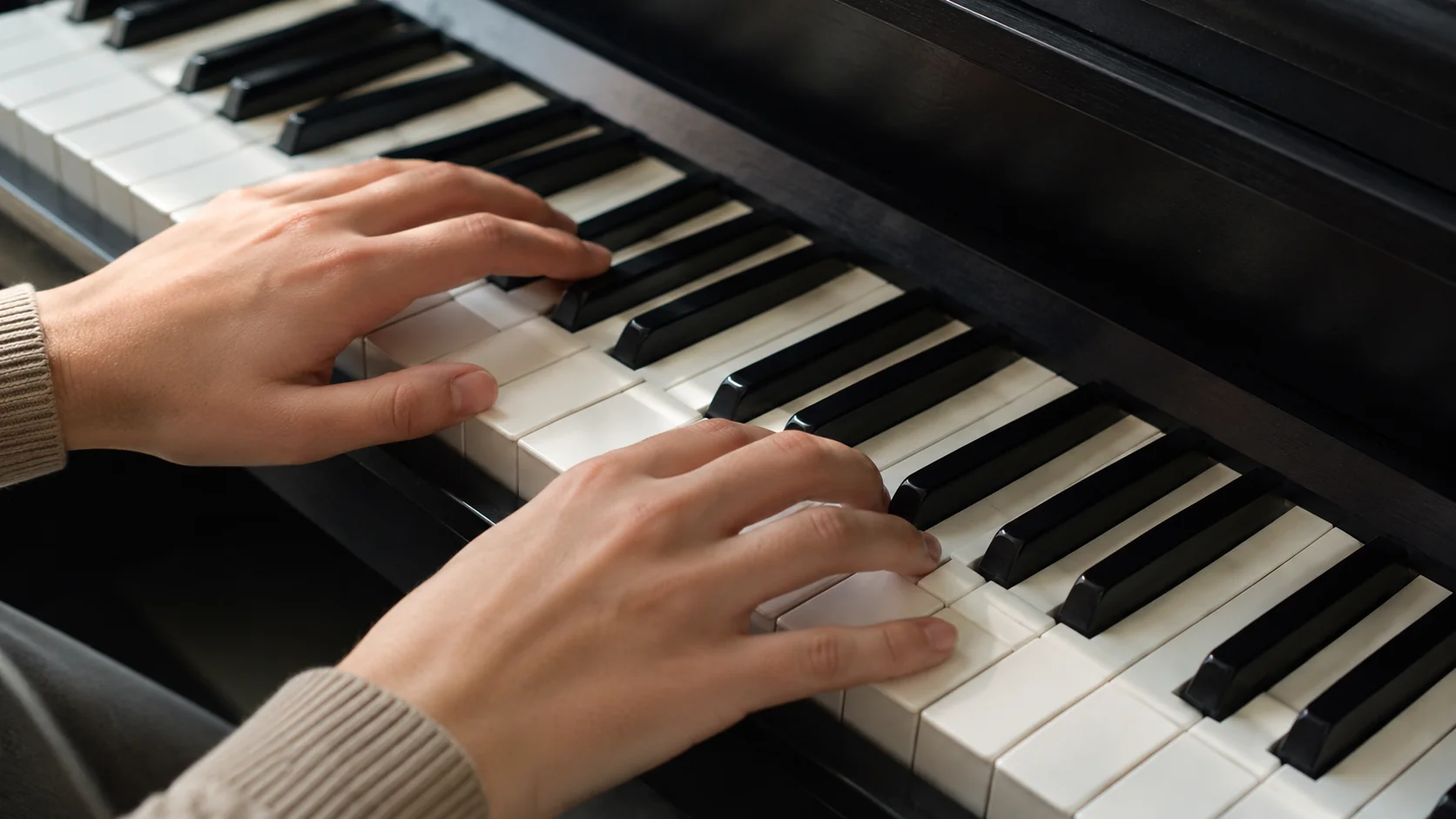 Vista próxima de duas mãos sobre teclas de piano em posição de iniciante, mão direita apoiada do polegar no dó central, luz suave de janela sobre o teclado