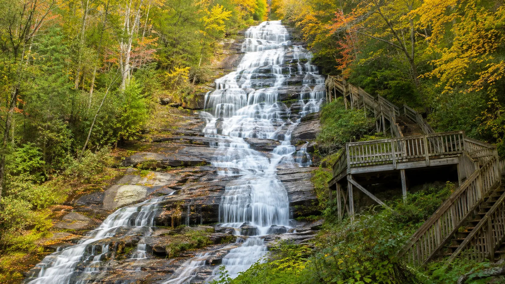 Escena ilustrativa: cascada alta de montaña bajando una ladera boscosa con escalera de madera y plataforma de observación al lado, típica de las cascadas de parque estatal del norte de Georgia