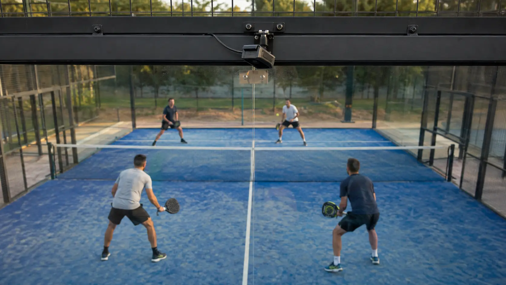 Cámara compacta montada sobre el vidrio del fondo de una cancha de pádel cerrada, jugadores visibles a través de él