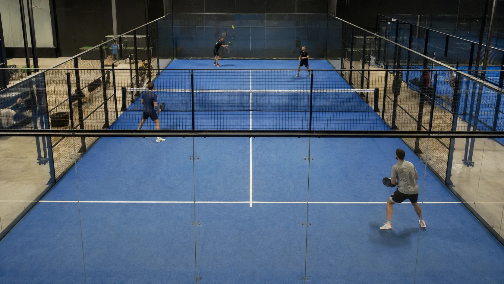 Two padel players seen from behind the back glass, one at the net and one recovering to the baseline during a rally