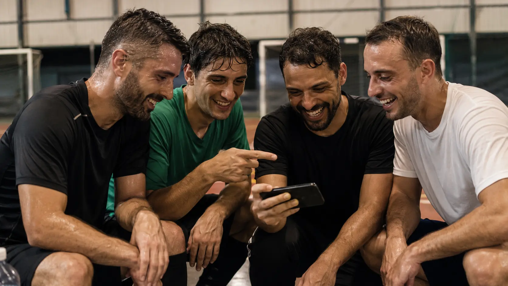 Amateur soccer players at the side of an indoor court celebrating while watching a goal clip on one phone