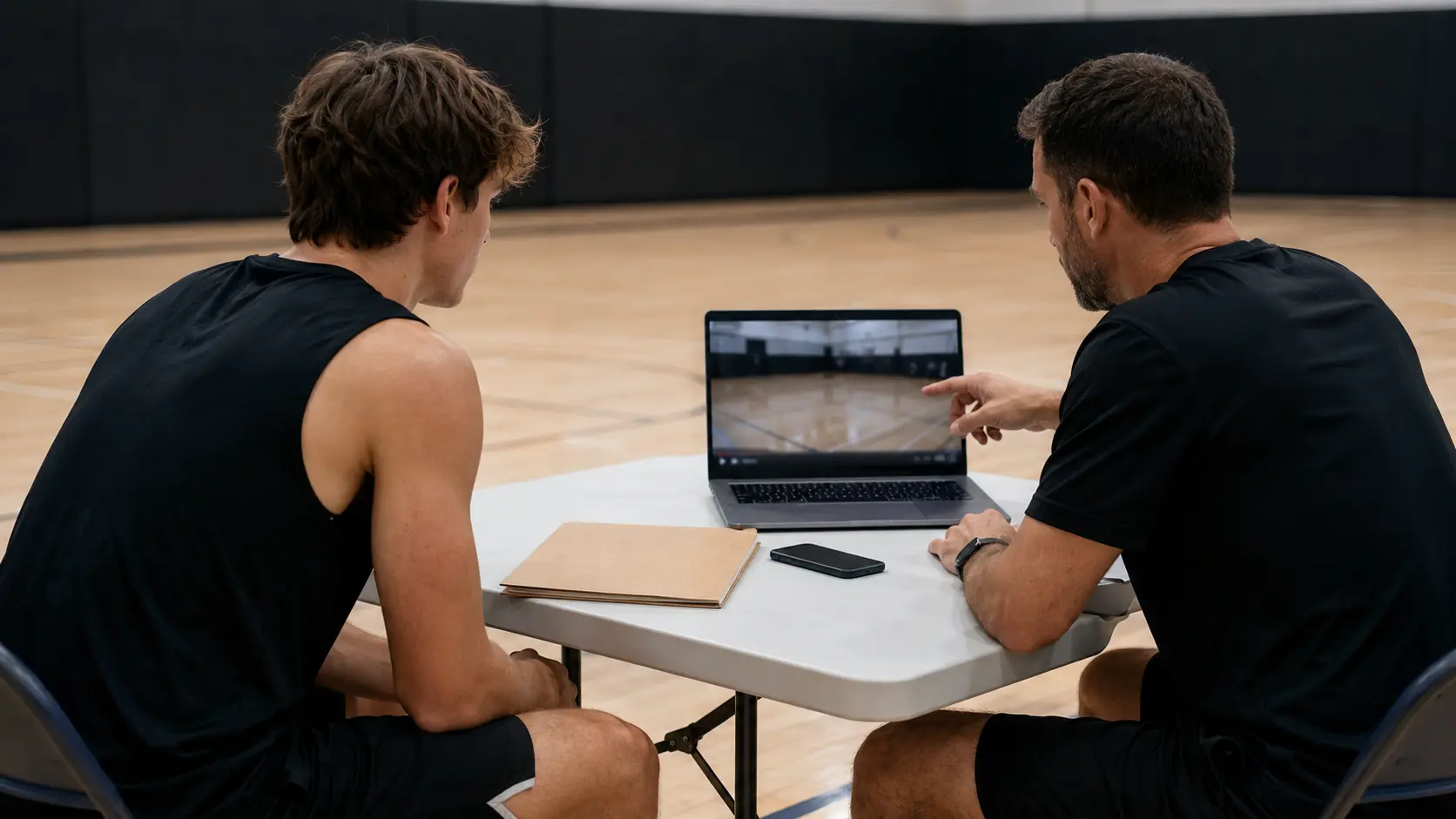Amateur athlete and coach reviewing a finished sports highlight reel on a laptop beside a court, with a phone and labeled source folder nearby and no recognizable software interface