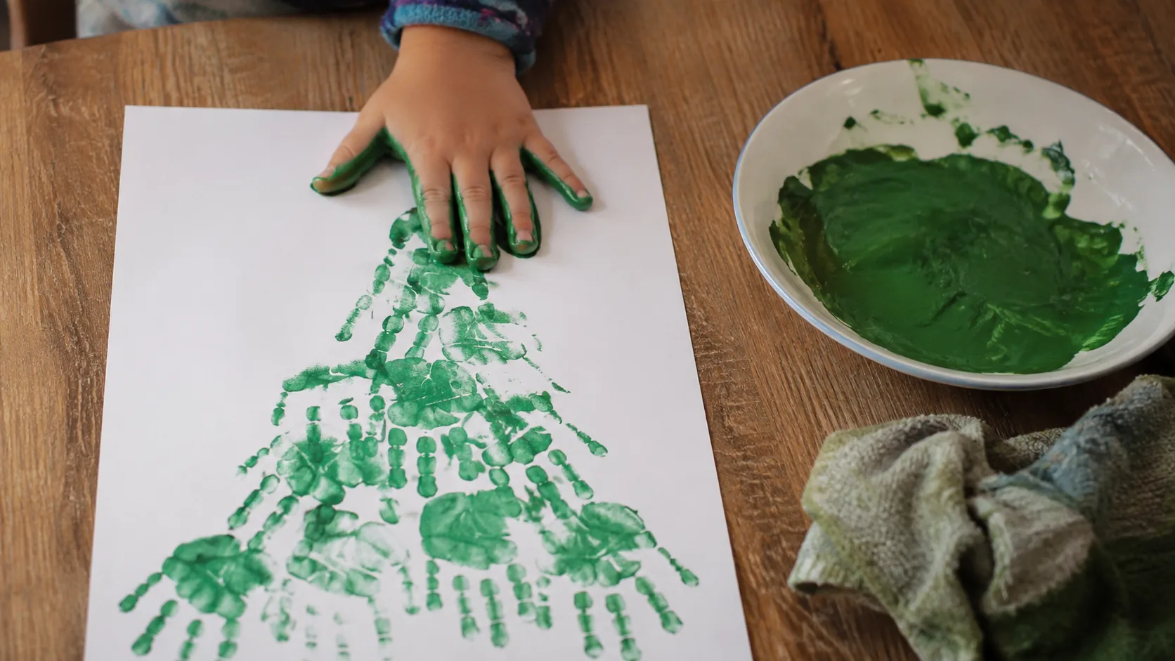 A small hand painted green being pressed onto a sheet of card at a low table, with a row of green handprints below it forming a tree shape and a plate of paint nearby