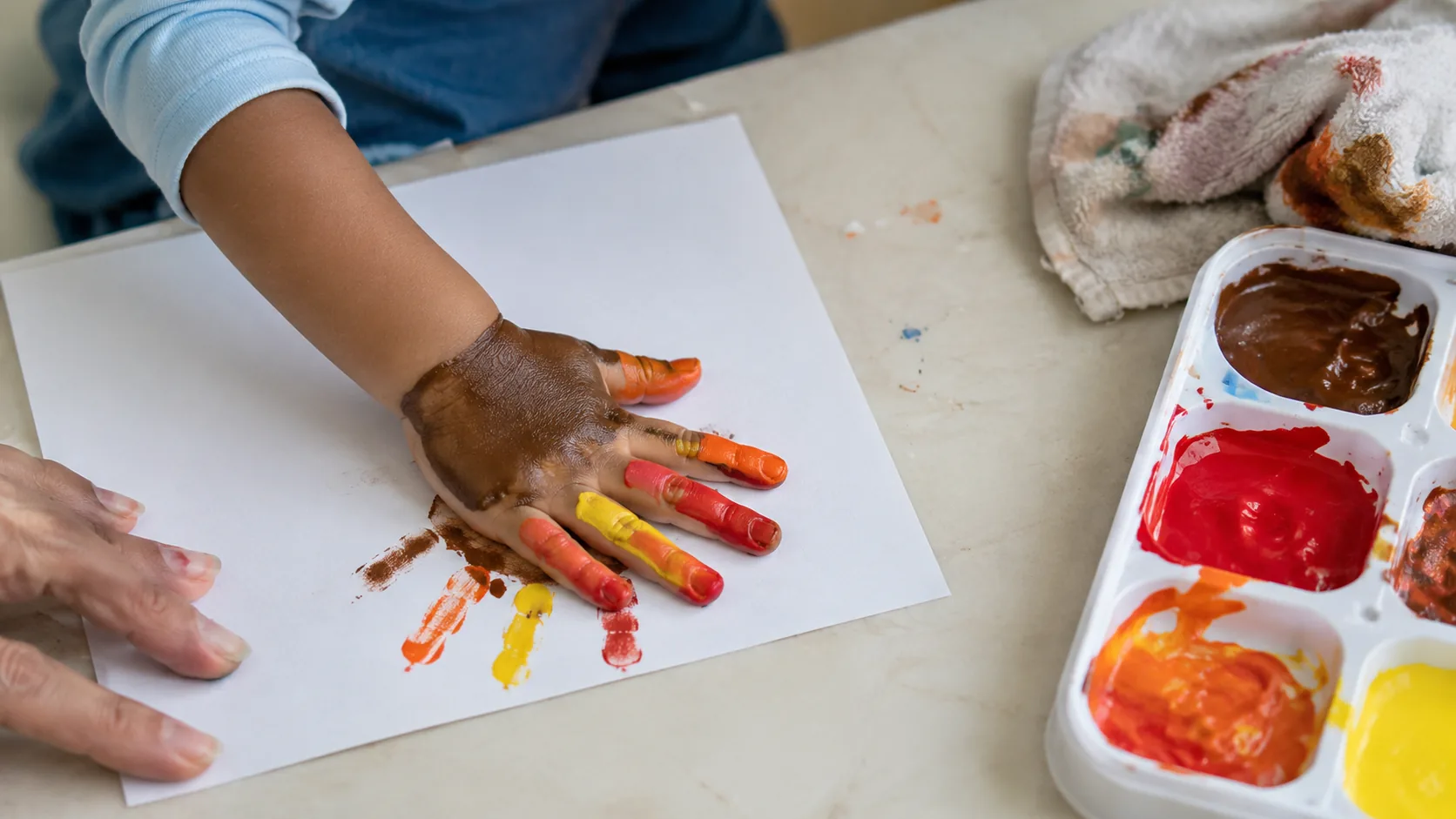 Mão de criança pequena pintada de marrom e laranja sendo apertada sobre cartolina branca numa mesa baixa, com uma bandeja de tinta lavável e um pano molhado ao lado