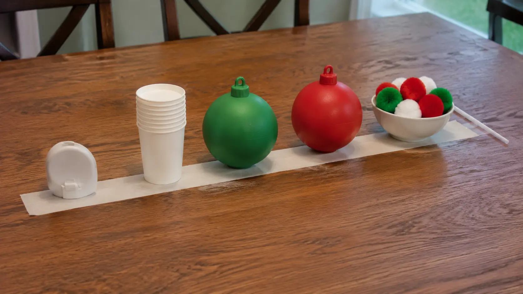 Mesa de comedor preparada para un reto de un minuto, con vasos de papel, bolas de Navidad de plástico, un cuenco de pompones grandes, una pajita y un temporizador de cocina sobre una tira de cinta de pintor