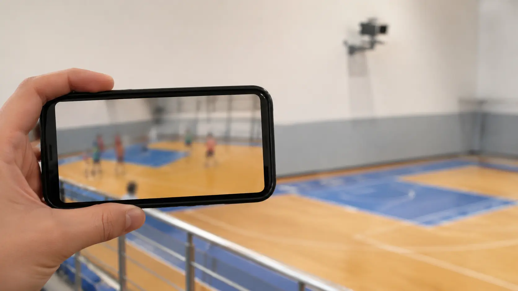 Celular en primer plano con la miniatura borrosa de un clip deportivo corto, y una cámara fija genérica instalada en lo alto de una cancha cubierta al fondo