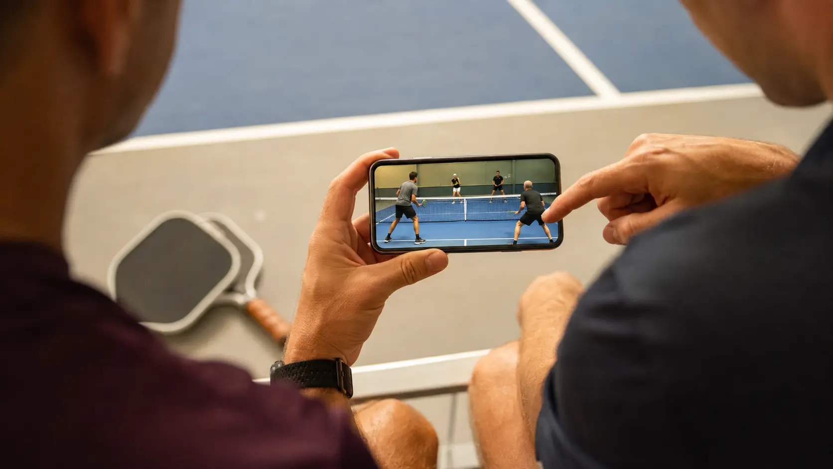Dupla de pickleball sentada depois do jogo olhando um clipe de rally em celular sem marca e sem texto legível na tela