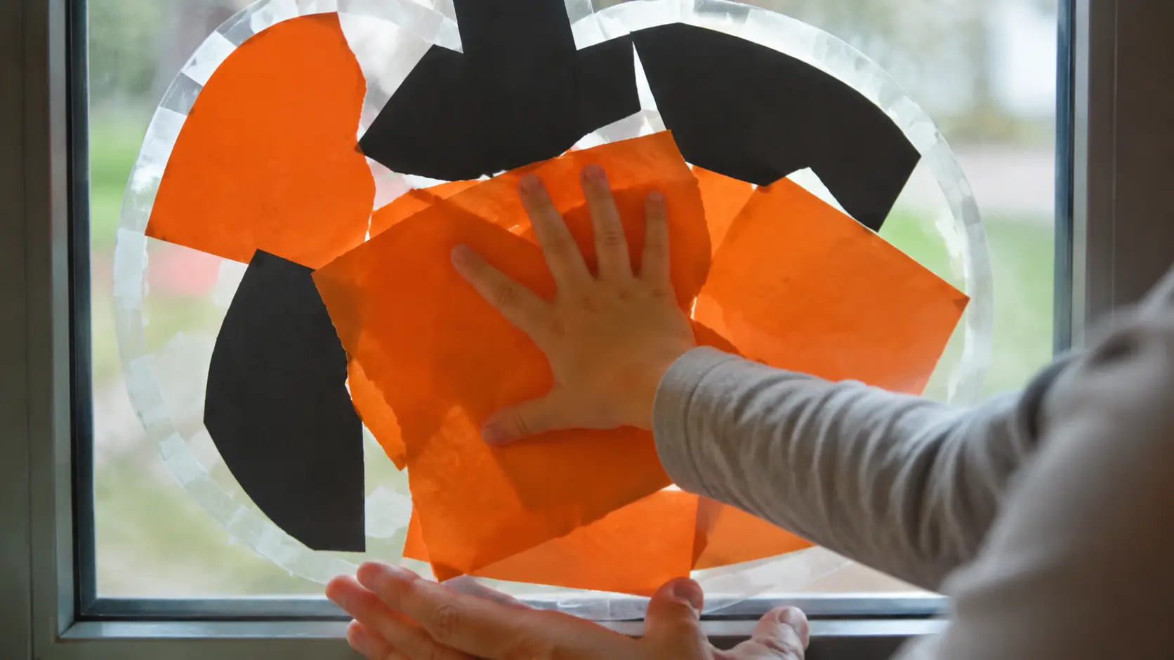 Mão pequena pressionando um pedaço grande de papel laranja rasgado numa abóbora redonda de papel adesivo transparente presa numa janela baixa, com alguns pedaços laranja e pretos já grudados e uma mão de adulto segurando a borda de baixo