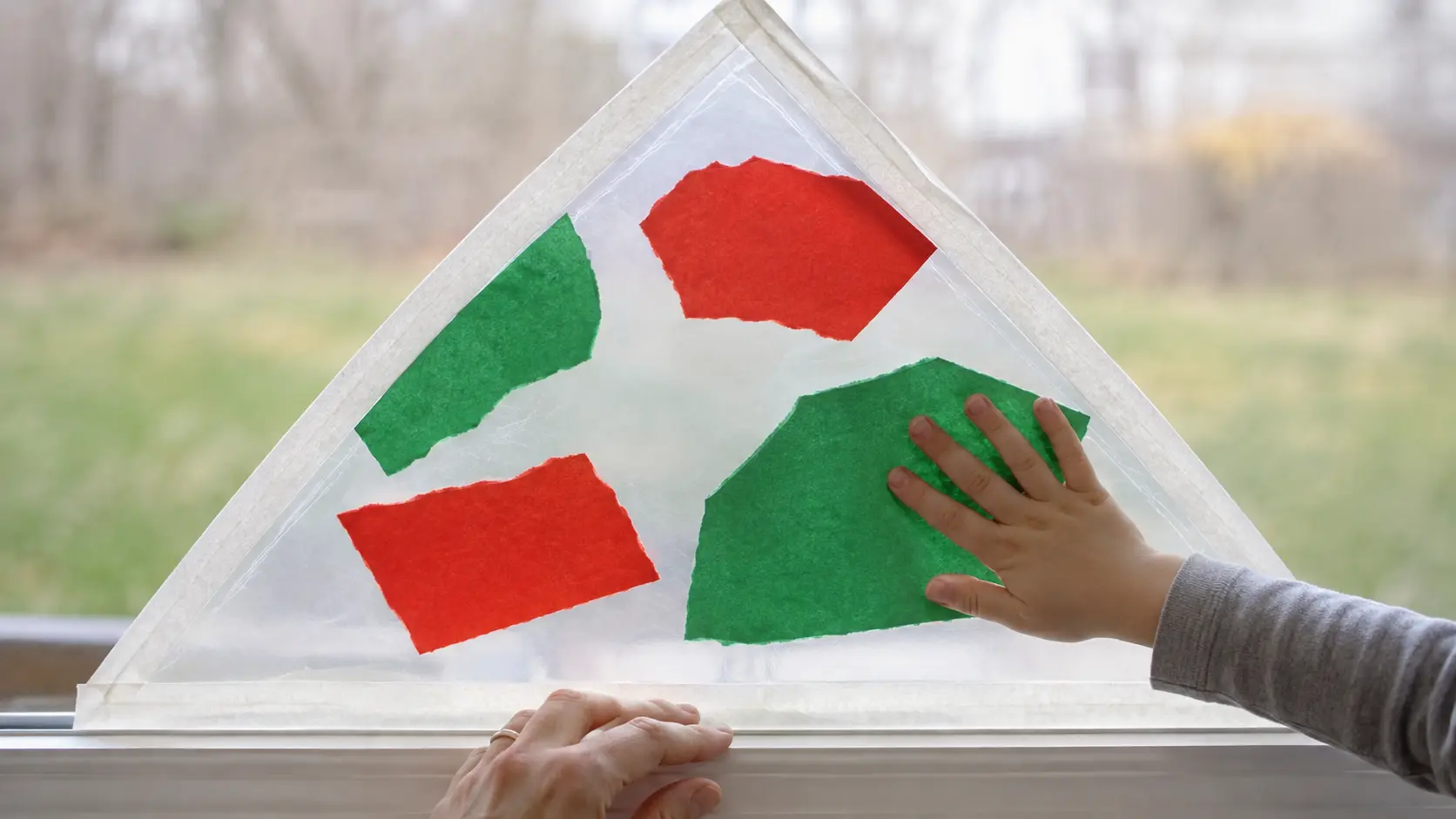 Mano pequeña presionando un trozo grande de papel verde rasgado sobre un triángulo de papel adhesivo transparente sujeto a una ventana baja, con algunos trozos rojos y verdes ya pegados y una mano de adulto sujetando el borde inferior