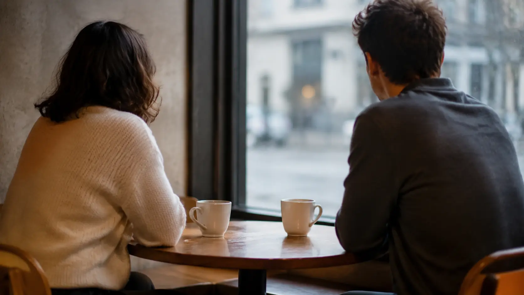 Dos personas vistas de lado en una mesa pequeña de cafetería junto a una ventana, dos tazas entre ellas, rostros girados lejos de la cámara