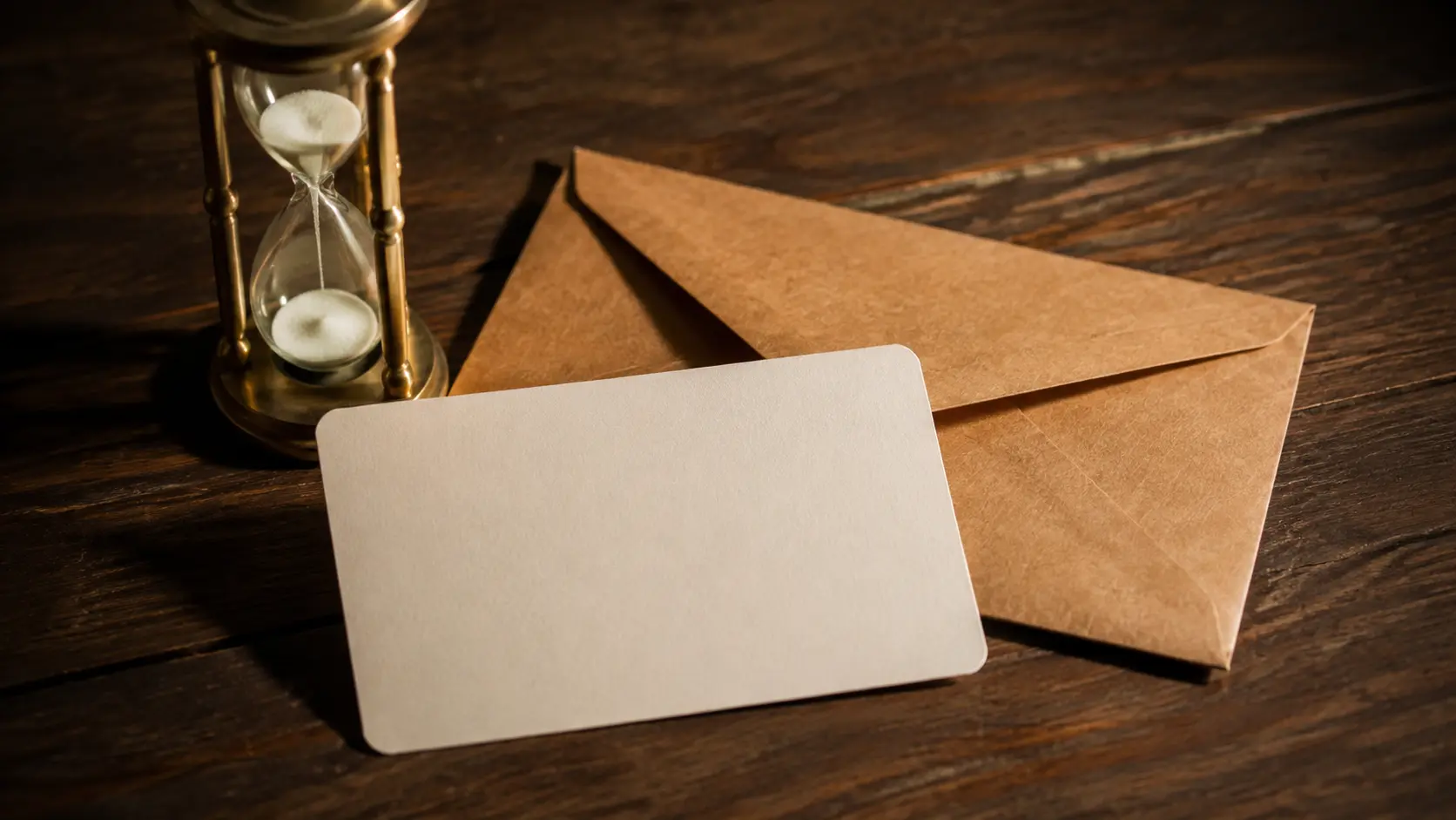 Cartão-presente sem marca, envelope fechado e pequena ampulheta organizados sobre mesa escura, sem logotipo nem texto legível