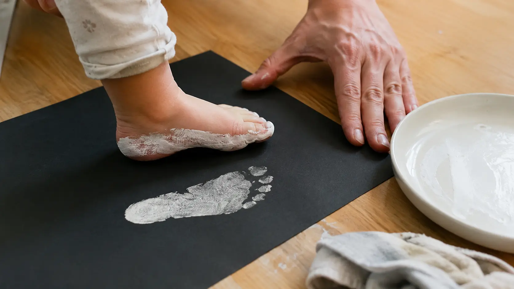 Mão de adulto segurando uma cartolina preta esticada numa mesa baixa enquanto um pé pequeno descalço pintado de branco é pressionado nela, com um prato de tinta branca e um pano úmido ao lado da cartolina